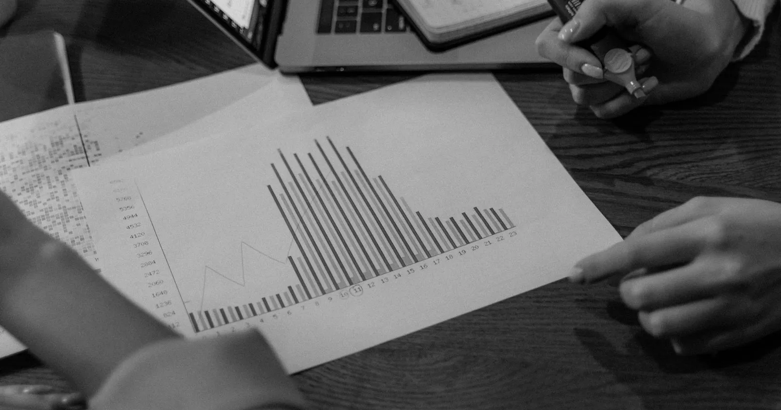 Two people reviewing a printed bar chart of website traffic on a wooden desk, next to a laptop and a notebook